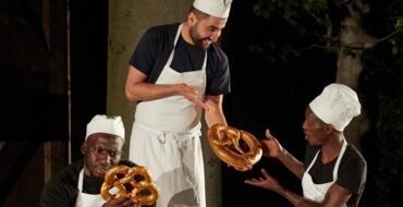 Hier sieht man einen jungen Mann, der in Bäckerkleidung in der Mitte steht und einem anderen jungen Mann in Bäckerkleidung auf einem Mehlsack sitzend eine Breze zeigt. Links sitzt auch ein junger Mann auf einem Mehlsack und sieht sich eine Breze an.