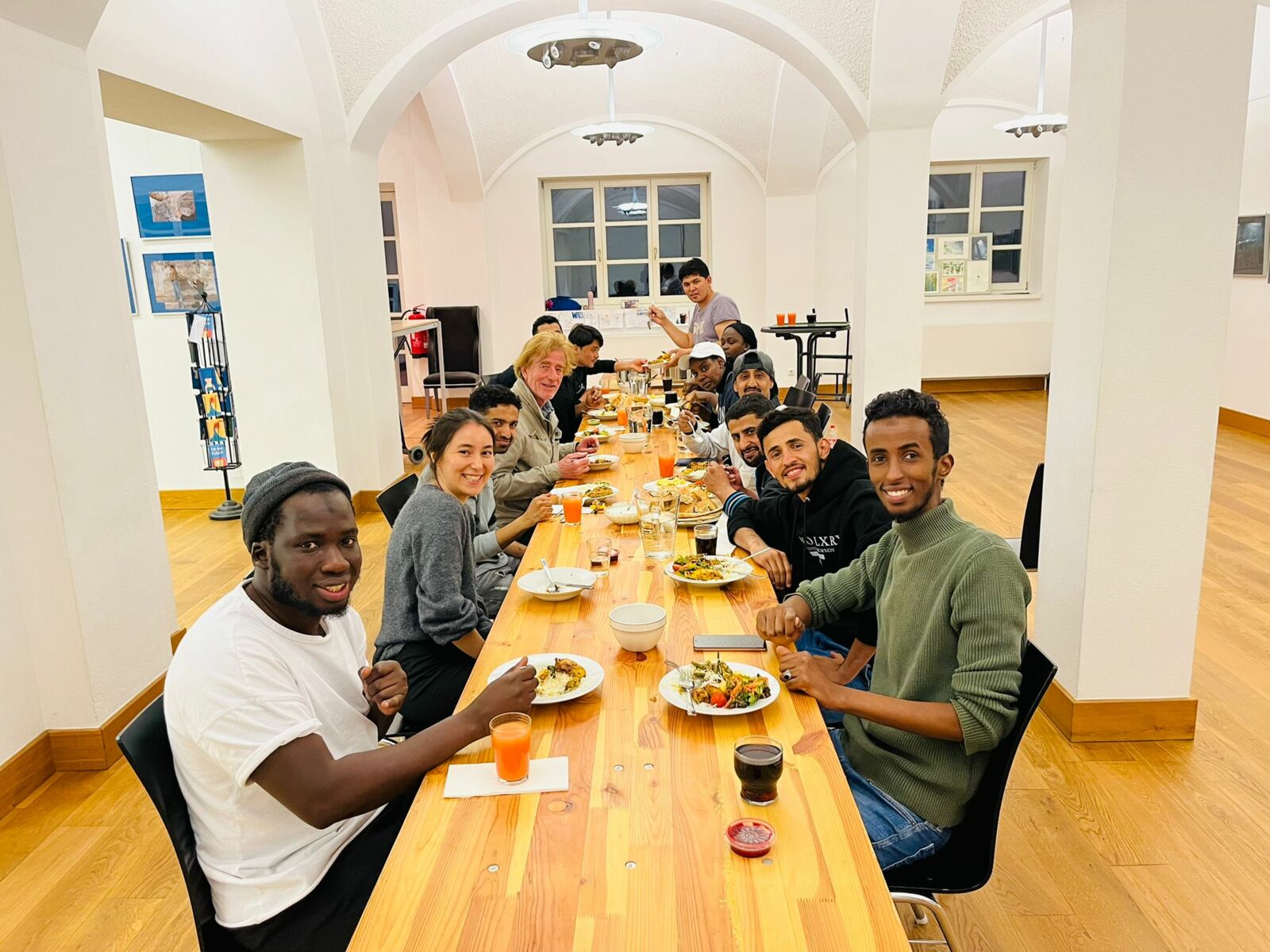 Hier sieht man ein Gruppenfoto von Menschen, die an einem langen Tisch mit Essen und Trinken sitzen und in die Kamera lachen.