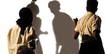 Hier sieht man zwei Schauspieler, die mit dem Rücken zum Publikum stehen. Sie blicken auf eine weiße Leinwand, auf der man die Schatten von zwei weiteren Menschen sieht, die gerade mit einem Selfiestick ein Foto machen.