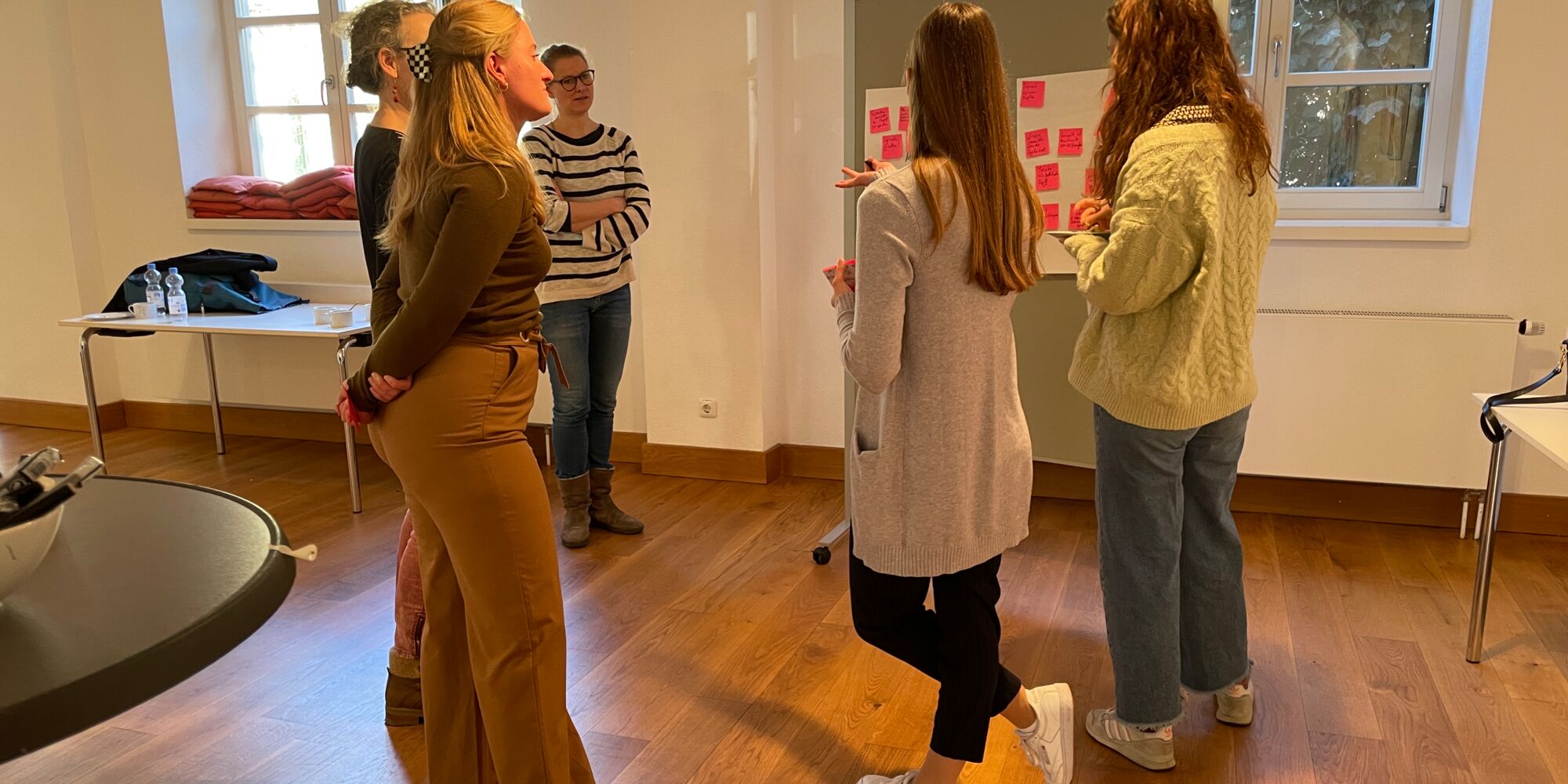 Hier sieht man mehrere Frauen, die im Halbkreis vor einem Whiteboard stehen und sich unterhalten.