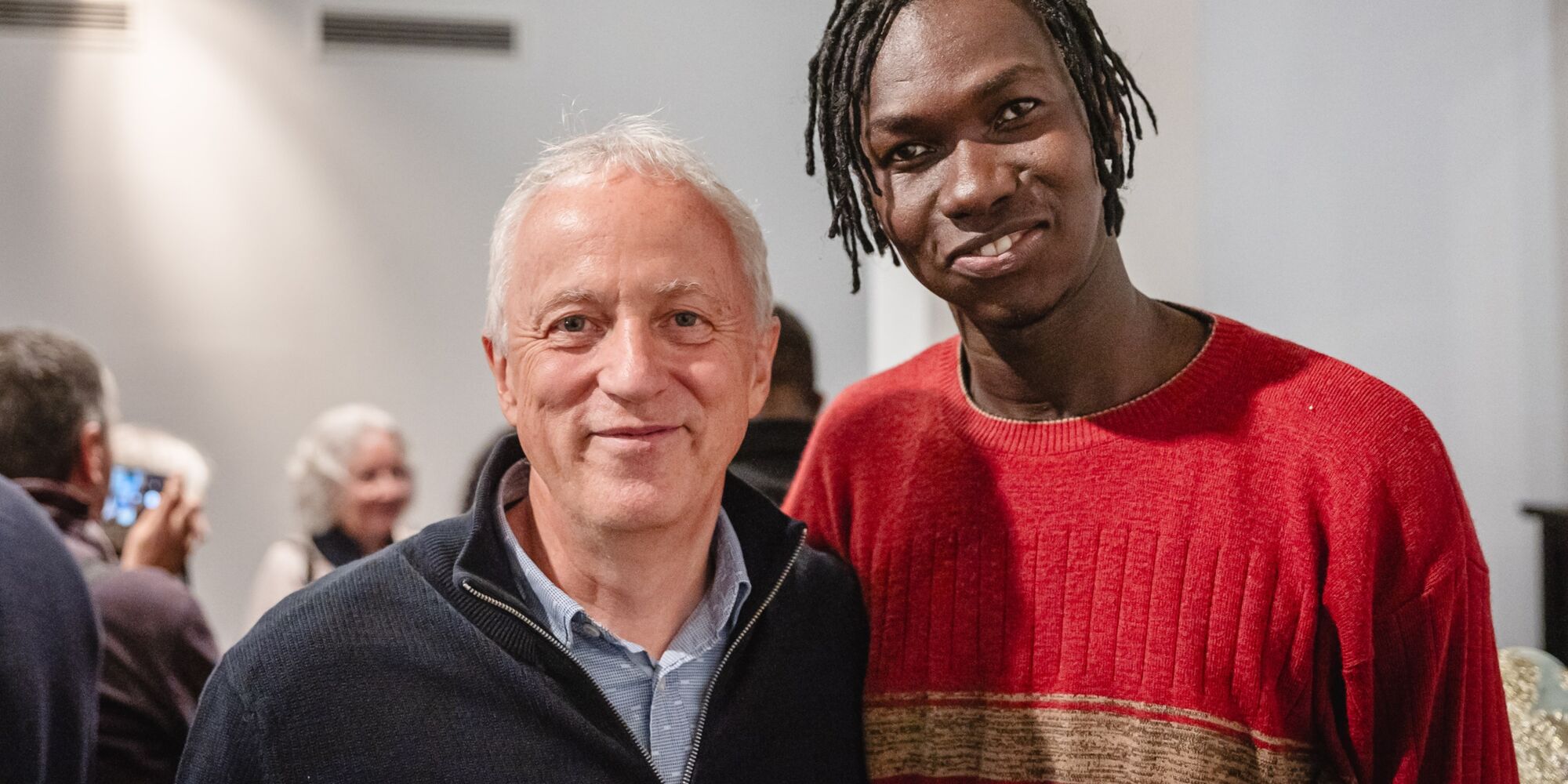 Hier sieht man einen älteren Herrn, wie er mit einem jungen Mann aus dem Ensemble für ein Foto posiert. Beide lächeln in die Kamera und halten sich gegenseitig am Rücken.