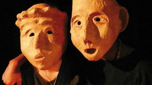 Hier sieht man zwei Schauspieler mit Masken, die ihre Gesichter komplett verdecken. Der eine hat den Arm um den anderen gelegt.