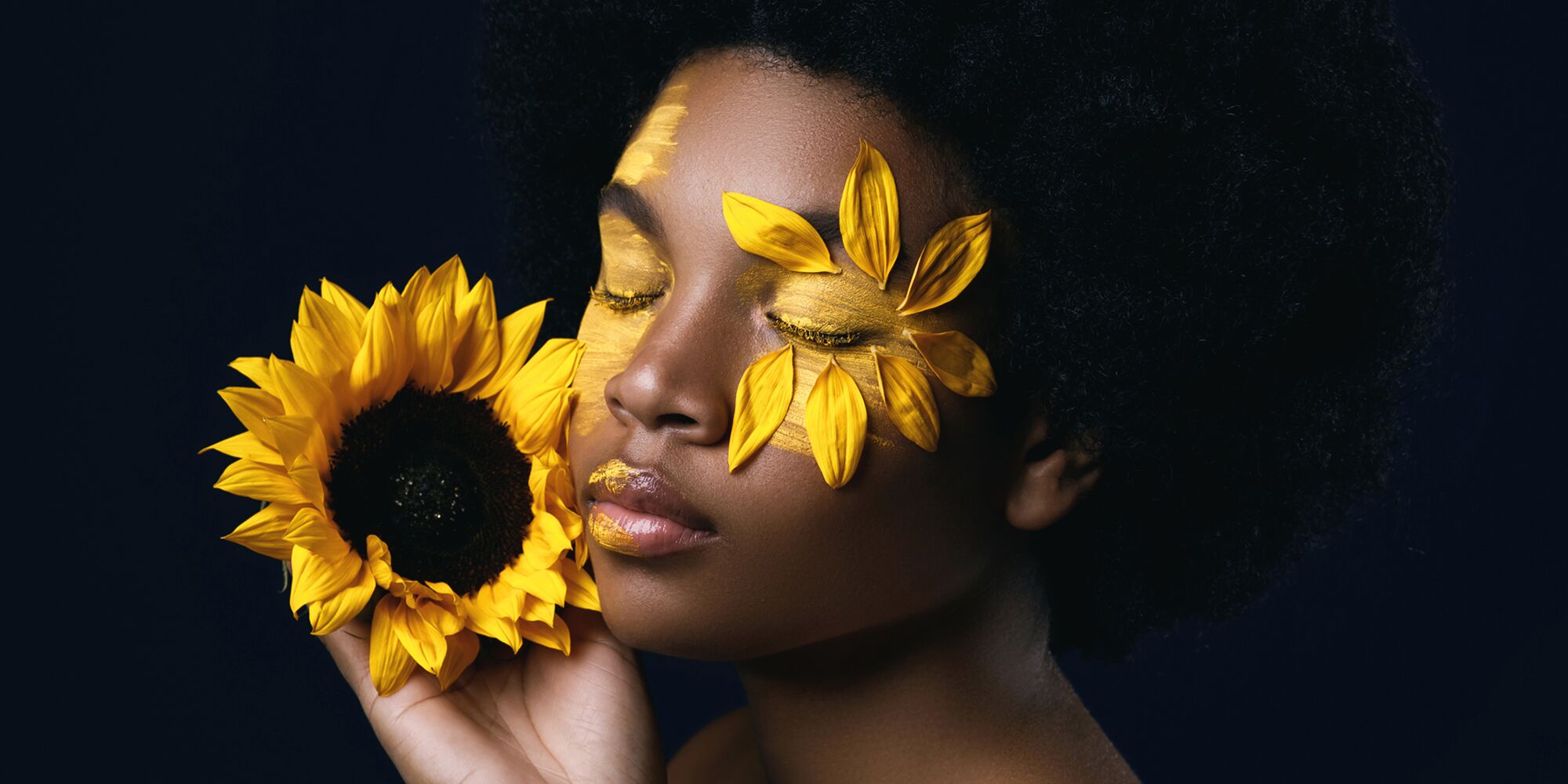 Hier sieht man eine Schwarze Frau, die die Augen zu hat und sich eine Sonnenblume neben ihr Gesicht hält. Ihre Augen sind gelb angemalt, um eines herum sind Sonnenblumenblüten.