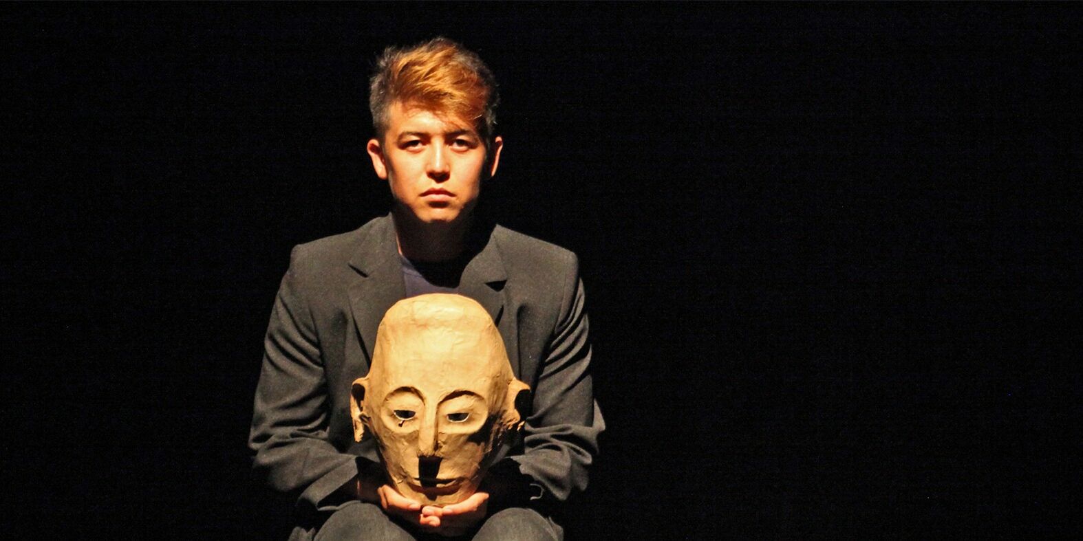 Hier sieht man einen jungen Schauspieler der sitzt. Der Hintergrund ist schwarz. Er sieht direkt in die Kamera und hält eine Maske in seinen Händen.