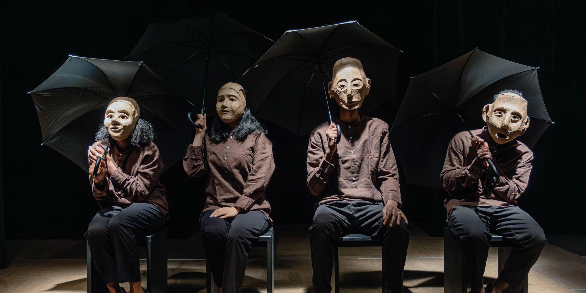 Hier sieht man vier Personen mit braunen Hemden und Masken, die ihre Gesichter komplett verdecken, nebeneinander auf Stühlen sitzen und einen jeweils einen schwarzen Regenschirm halten.