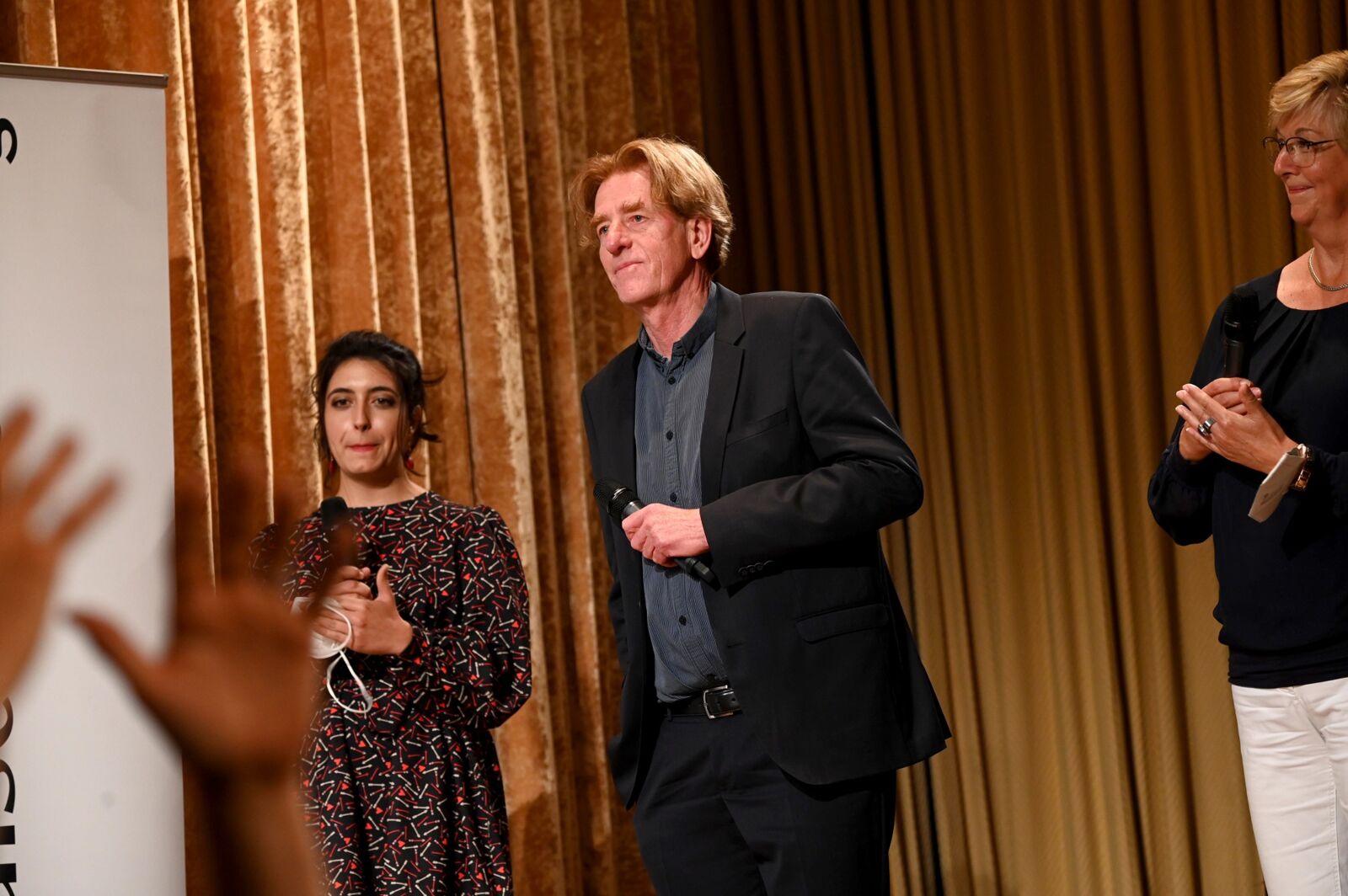 Hier sieht man Viktor Schenkel mit einem Mikrofon in der Hand auf der Preisverleihung des Tassilo-Sozialpreises 2021, links von ihm Sophie Pacini. Im Vordergrund sind zwei Hände zu sehen.