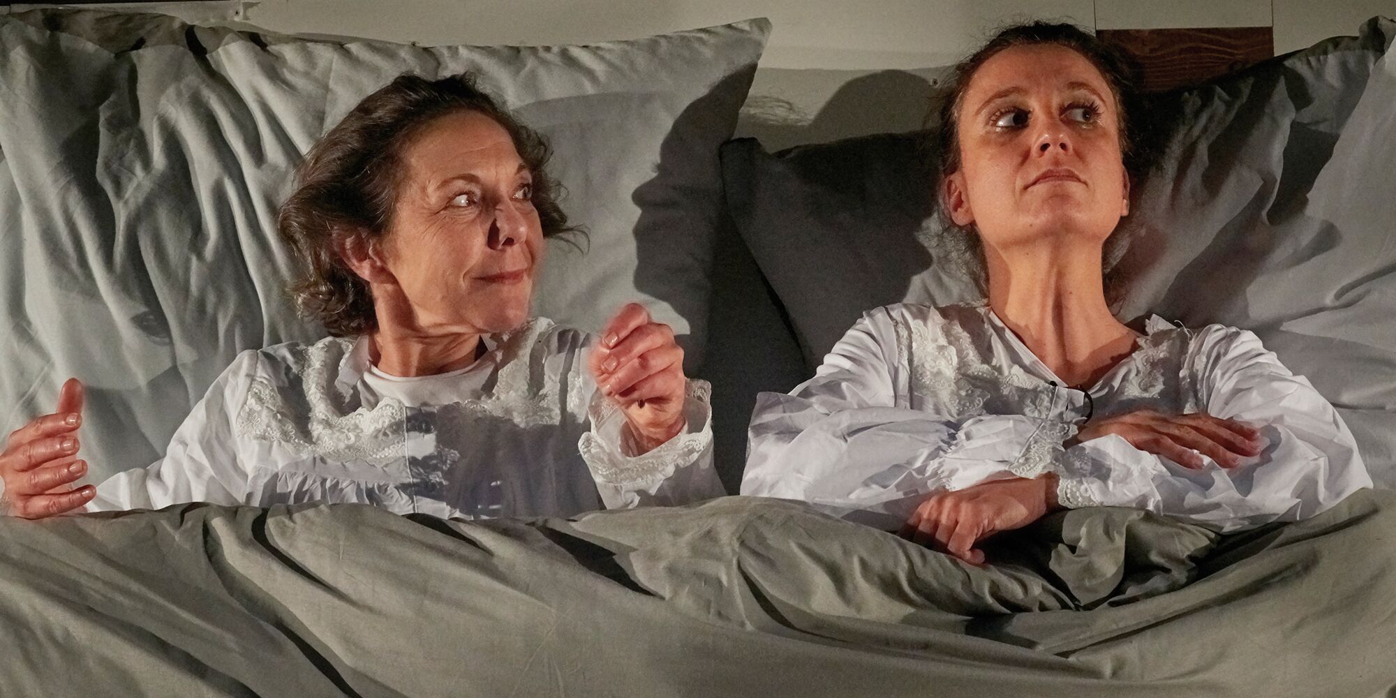 Zwei Frauen liegen im Nachthemd in einem Bett. Die Frau links blickt auf die andere Dame, diese sieht weg.