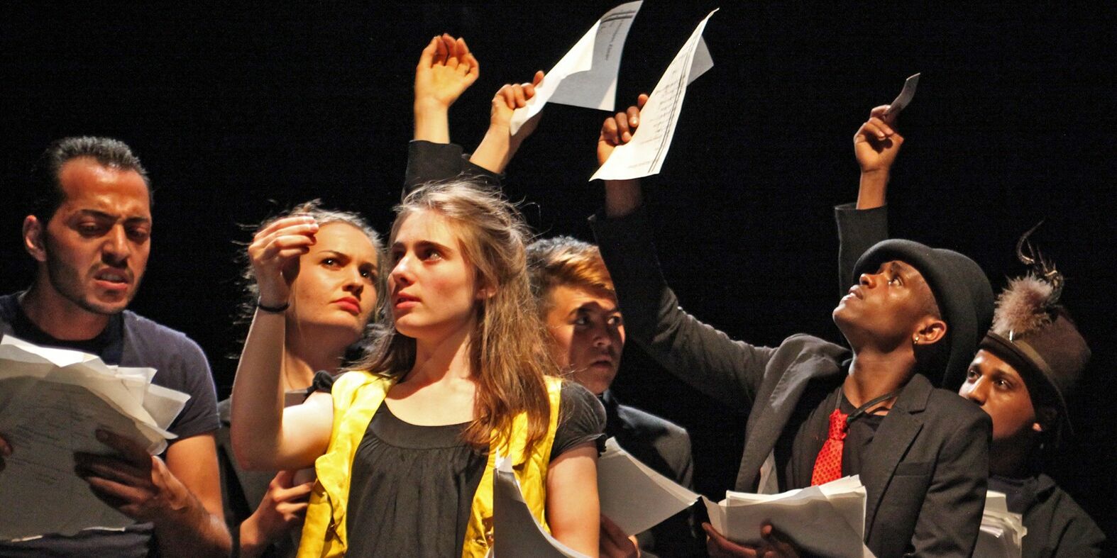 Hier sieht man mehrere Schauspieler und Schauspielerinnen des Stückes "Unterm Sprung" wobei viele Blätter hochhalten und diese ansehen. Ganz links hält einer einen Stapel dieser Blätter in der Hand udn schaut diesen wütend an.
