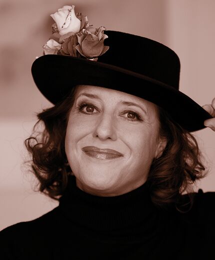 Hier sieht man ein Foto der Kabarettistin Luise Kinseher, die in die Kamera sieht und lächelt. Sie hat einen schwarzen Hut auf, den sie mit einer Hand festhält. Das Bild ist in Sepiatönen gehalten.
