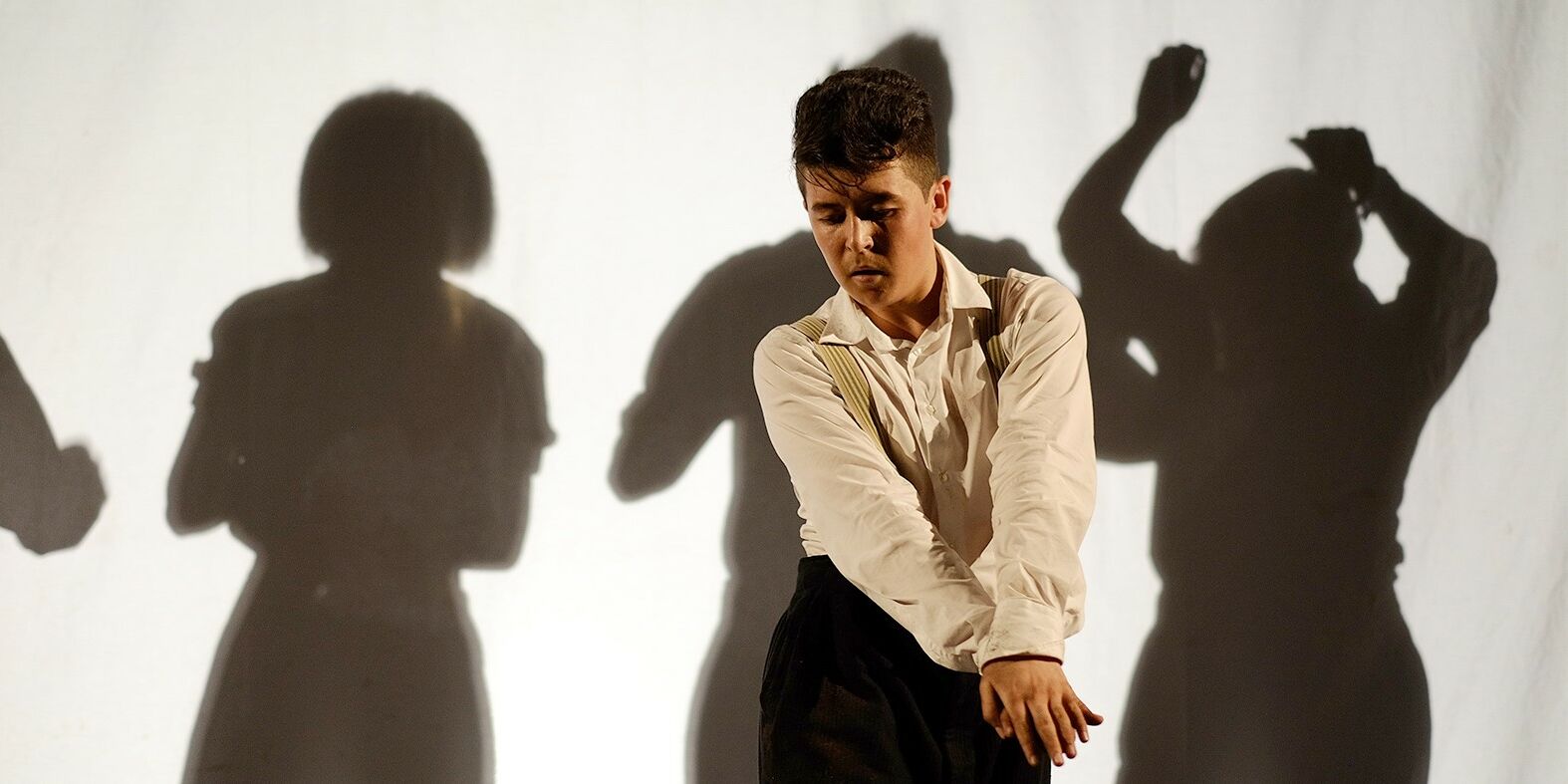 Hier sieht man ein Szenenfoto aus dem Stücke "Grenzenlos. Im Hintergrudn sieht man die Schatten mehrerer Personen. Vorne steht ein Schauspieler der die Hände aufeinandergelgt und vor sich ausgestreckt hat. Er sieht nach unten und hat den Kopf geneigt.