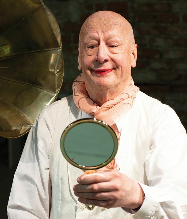 Ein älterer Mann mit Glatze, weißem Hemd, einem Seidentuch und rotem Lippenstift schaut in einen Spiegel. Auf der linken Seite sieht man ein Grammophon.