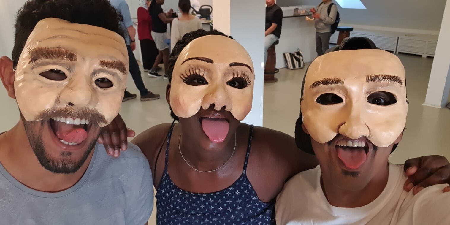 Hier sieht man drei Menschen, die für ein Gruppenselfie posieren. Sie alle tragen Masken und strecken die Zunge heraus.