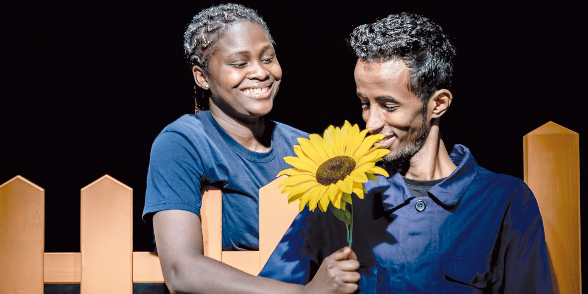 Hier sieht man links eine junge Frau, die einem jungen Mann rechts lächelnd eine Sonnenblume über einen Zaun reicht. Der junge Mann sieht die Blume an. Beide tragen dunkelblau und sind schwarz.