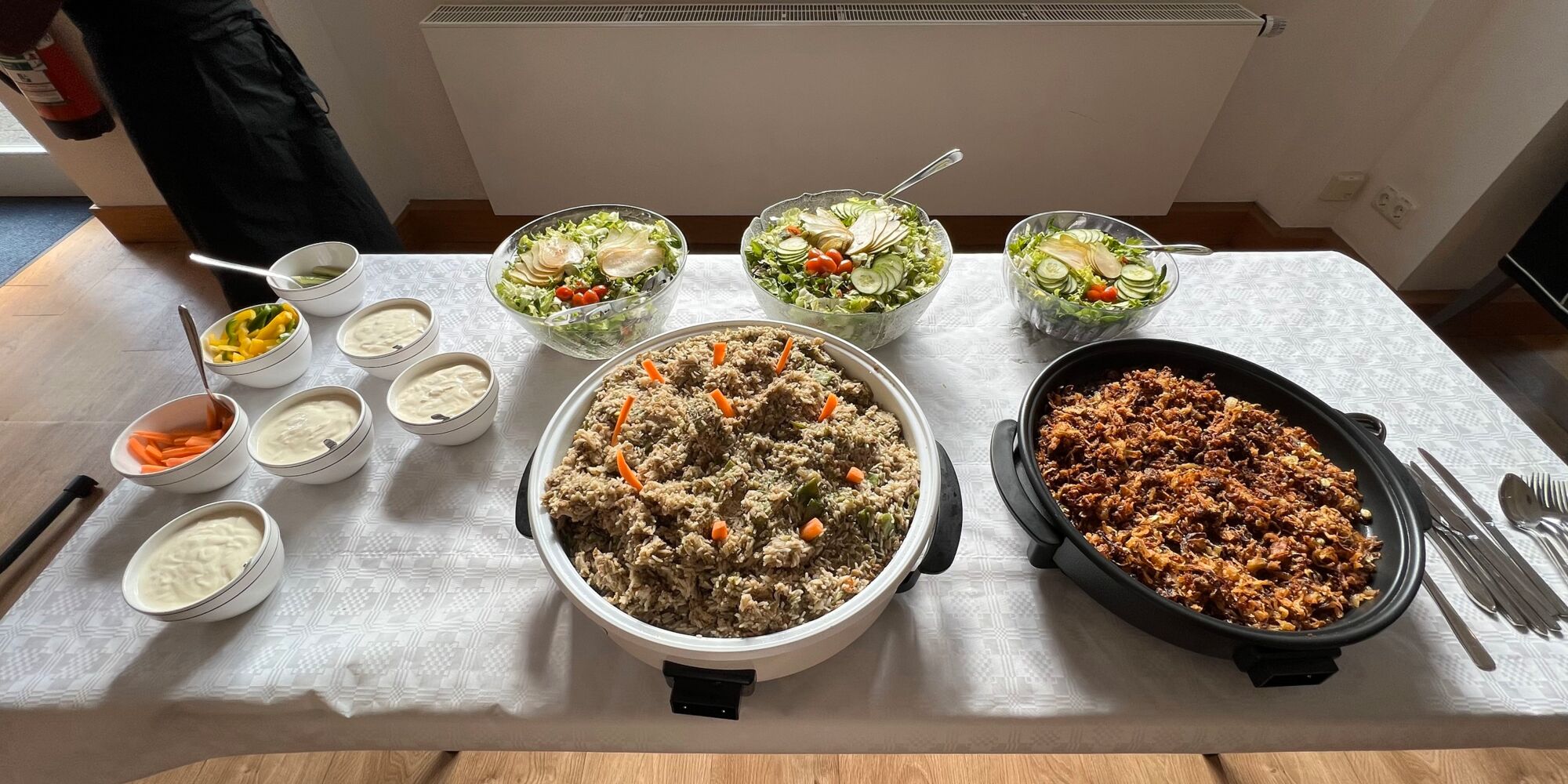 Hier sieht man mehrere Speisen in Schüsseln und Warmhaltepfannen, unter anderem drei Salate und mehrere Dips.