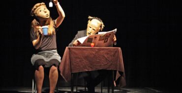 Hier sieht man zwei sitzende SchauspielerInnen, die Masken tragen. Die auf der linken Seite hält eine Tasse in der Hand, in die sie gerade einen Teebeutel tunkt. Der rechte sitzt an einem Tisch mit einer Zeitung in der Hand und sieht in ihre Richtung.