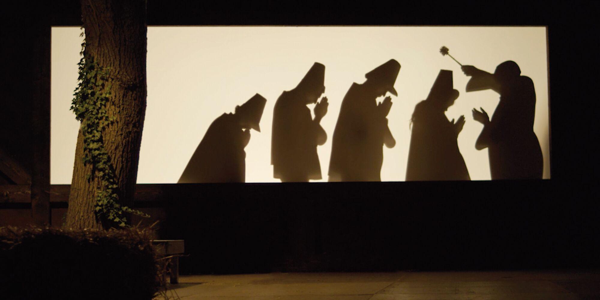 Man sieht eine Szene im Schattentheater. Ein Pfarrer segnet vier betende Silhouetten mit Weihwasser. Links im Vordergrund ein Baum zu sehen.