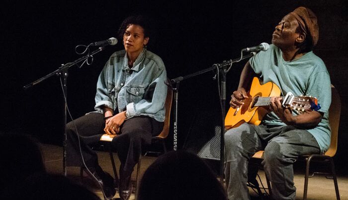 Hier sieht man das Vater-Tochter-Duo Wally und Ami Warning, die auf Stühlen auf der Bühne sitzen und singen. Wally Warning, der rechts sitzt, spielt dazu Gitarre.