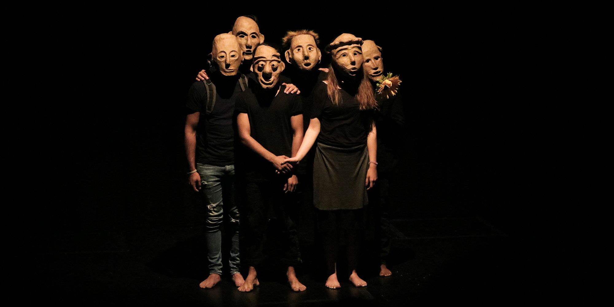 Hier sieht man Schauspielerinnen und Schauspieler mit Masken, die ihr komplettes Gesicht verdecken, in zwei Reihen dicht beieinander stehen. Sie sehen alle gerade nach vorne.