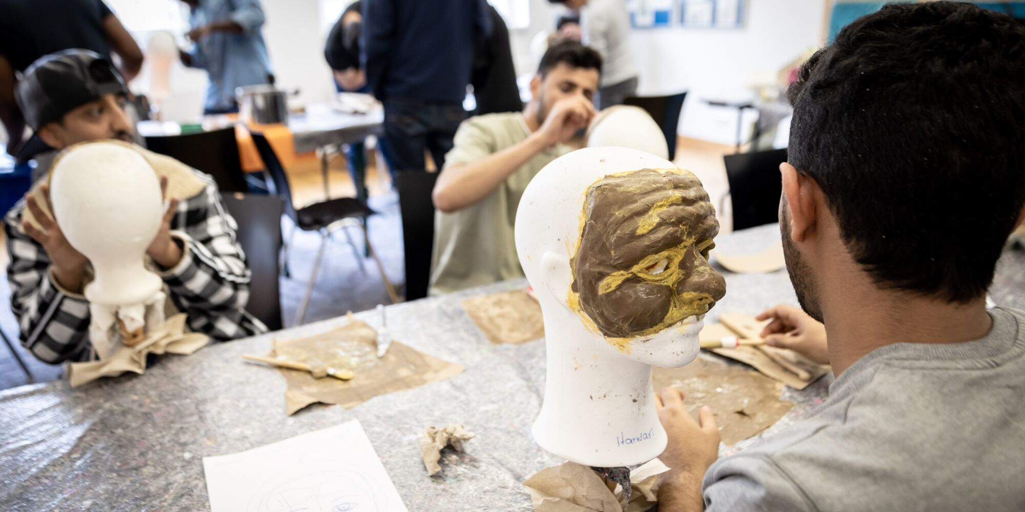 Hier sieht man drei junge Männer an einem Tisch, die an Masken auf Modellköpfen arbeiten. Im Hintergrund sind andere Menschen an ihren Masken arbeitend zu sehen.