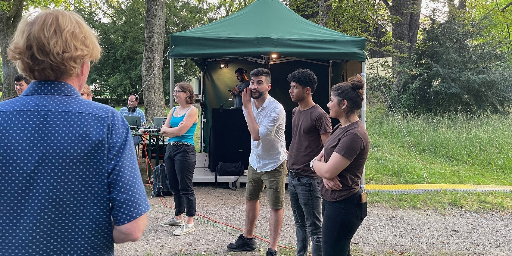 Hier sieht man mehrere Ensemblemitglieder von Theater Grenzenlos bei einer Teambesprechung im Park der Mohr-Villa. Im Hintergrund ist ein Technikzelt aufgebaut.