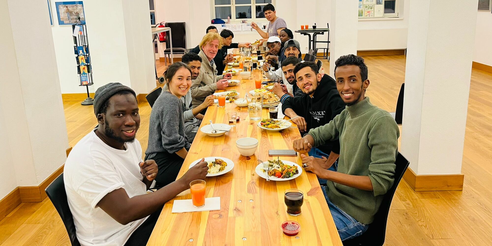 Hier sieht man ein Gruppenfoto von Menschen, die an einem langen Tisch mit Essen und Trinken sitzen und in die Kamera lachen.