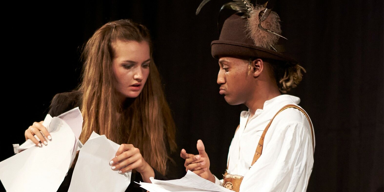 Hier sieht man eine Schauspielerin und einen Schauspieler. Die Schauspielerin hat mehrere Blätter in der Hand und sieht auf die Blätter die der andere in der hand hält. Dieser trägt bayerische Tracht und gestikuliert mit einer Hand.