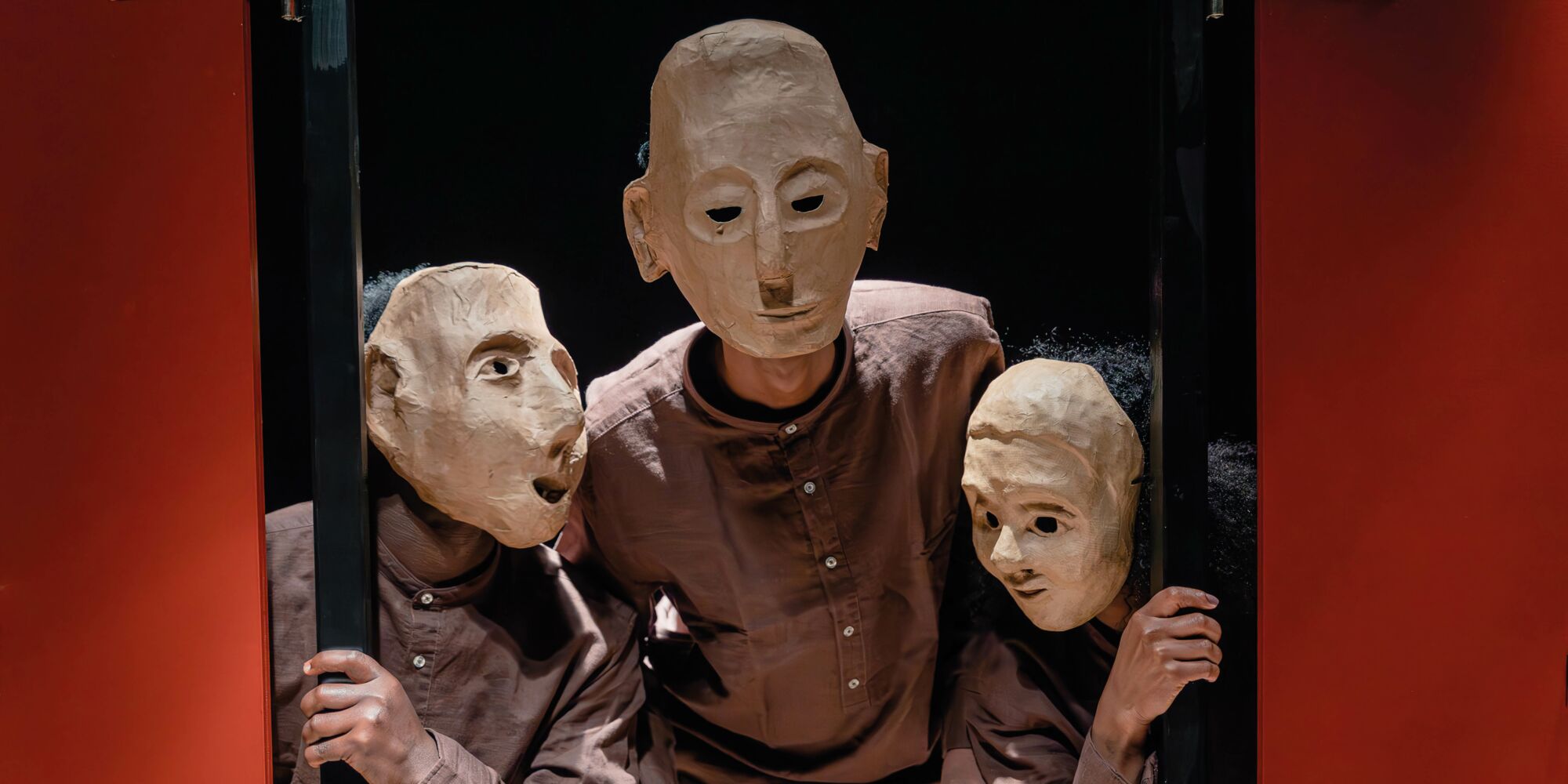Hier sieht man drei Personen mit braunen Hemden und Masken, die ihre Gesichter komplett verdecken, hinter roten Fensterläden hervorspähend.