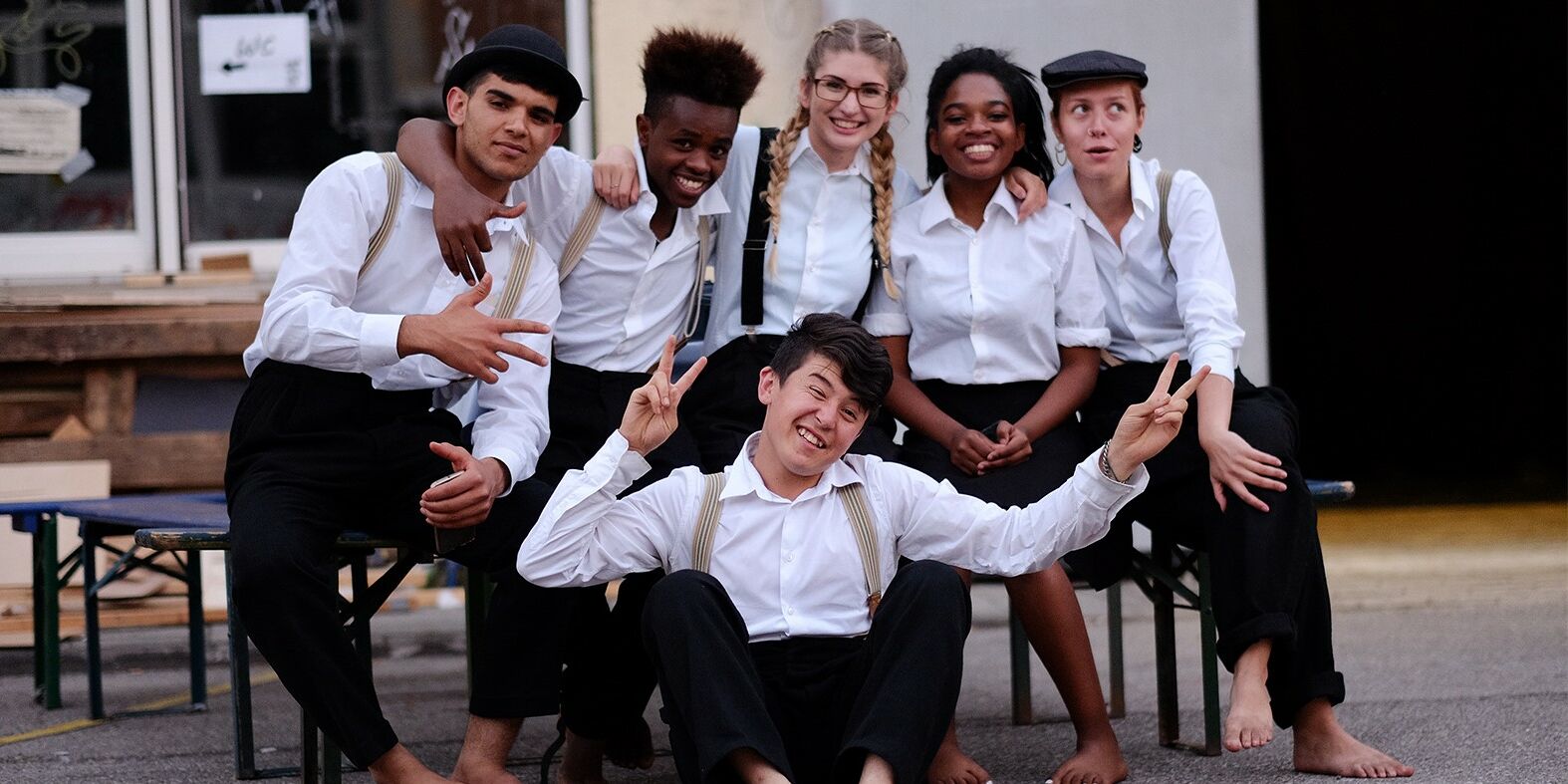 Auf disem Bild sind Schauspieler und Schauspielerinnen des Theater Grenzenlos zu sehen. Fünf sitzen auf einer Bank und haben die Arme umeinander gelegt. Vorne sitzt einer auf dem Boden, lacht und macht das Peace zeichen.