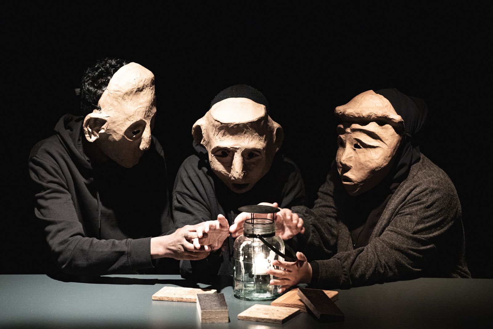 Hier sieht man drei Darsteller unseres Benefizabends 2022. Sie sitzen, tragen große Masken und wärmen sich ihre Hände an einer Laterne.