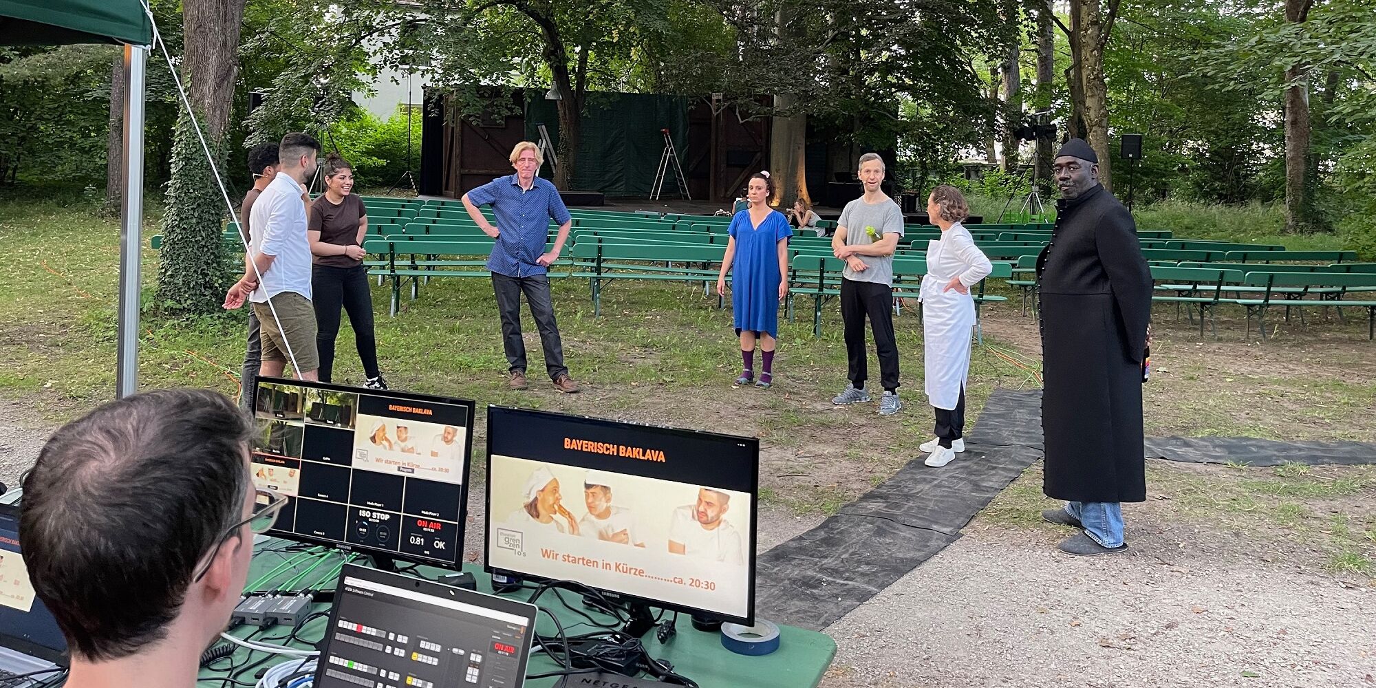 Hier sieht man mehrere Ensemblemitglieder von Theater Grenzenlos bei einer Teambesprechung im Park der Mohr-Villa. Im Hintergrund sieht man die Naturbühne mit den Sitzbänken für das Publikum, links im Vordergrund einen Techniker vor zwei Computern.