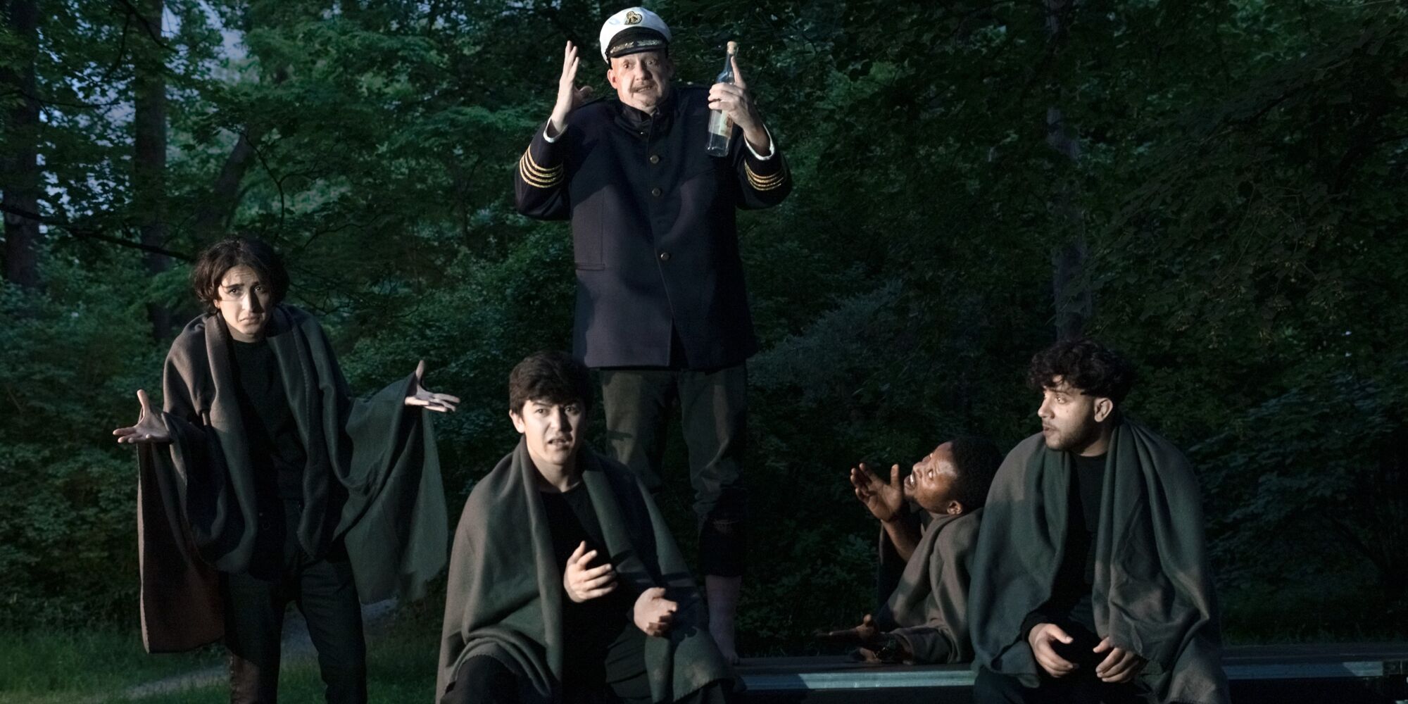 Vier junge Männer in schwarzen Klamotten und je einer dunklen Decke um die Schultern sitzen und stehen mit verwirrten Gesichtern um einen entsetzt aussehenden Schiffskapitän mit einer Schnapsflasche in der Hand.