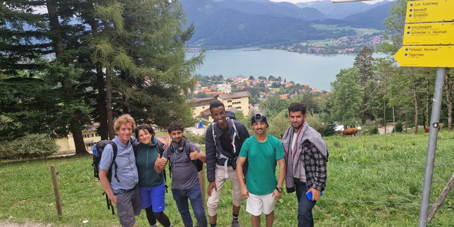 Hier sieht man ein Gruppenbild von wandernden Personen vor einem See. Rechts sieht man gelbe Wegweiser.