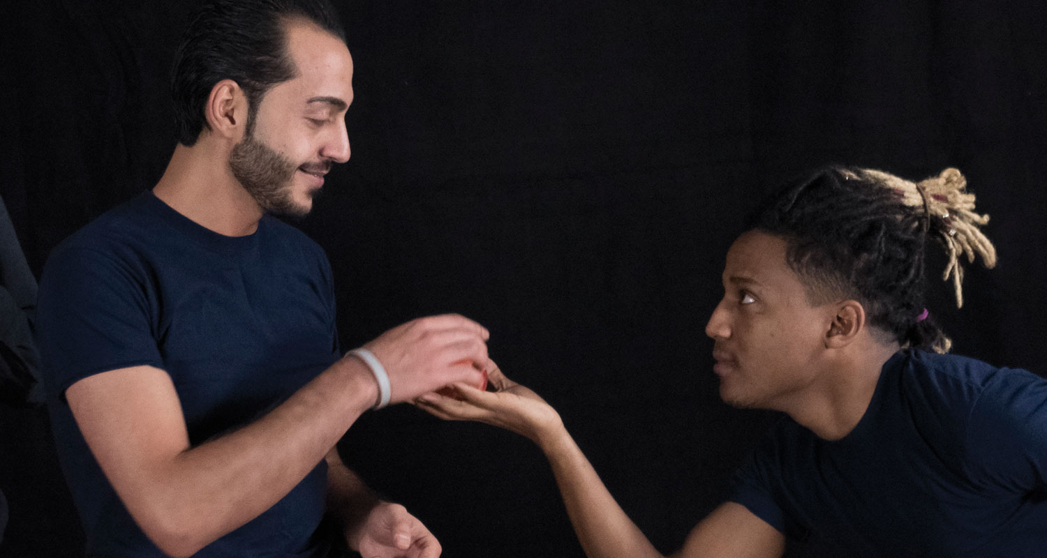 Zwei Schauspieler proben eine Szene, bei der der rechte Schauspieler dem linken einen Gegenstand auf seiner Hand anbietet. Dieser nimmt es gerade entgegen.