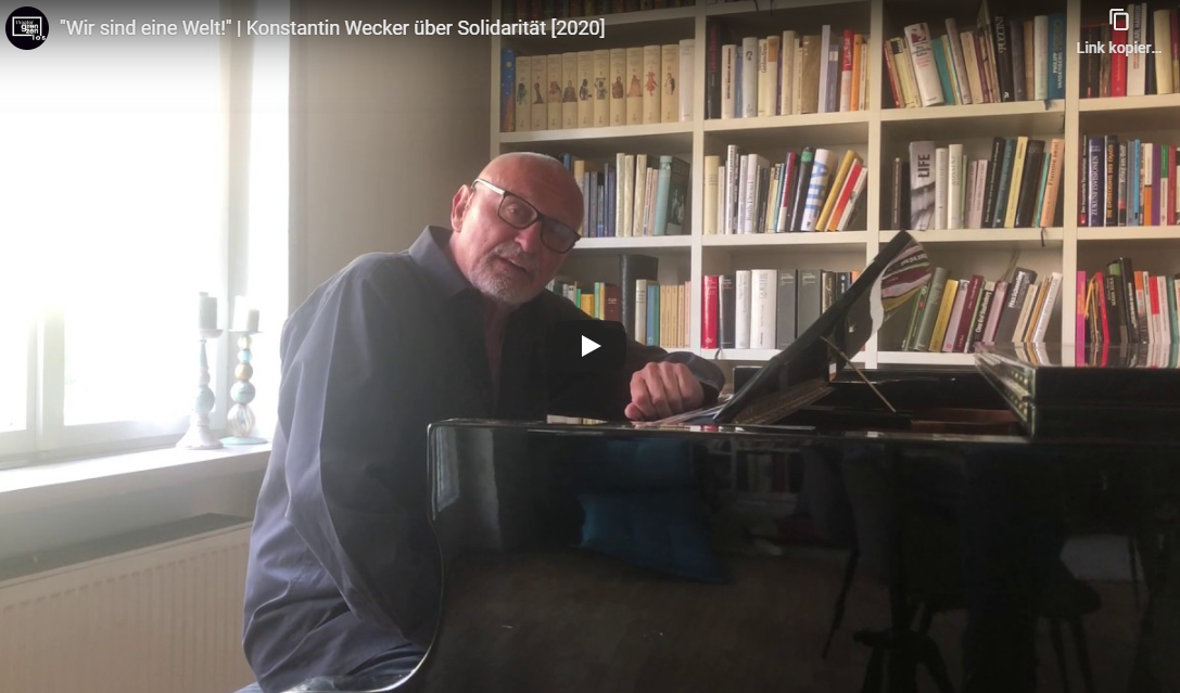 Hier sieht man einen Ausschnitt des Youtube-Videos "Wir sind eine Welt" von Konstantin Wecker. Dieser sitzt an einem Flügel und sieht in die Kamera. Im Hintergrund sieht man ein großes Bücherregal.