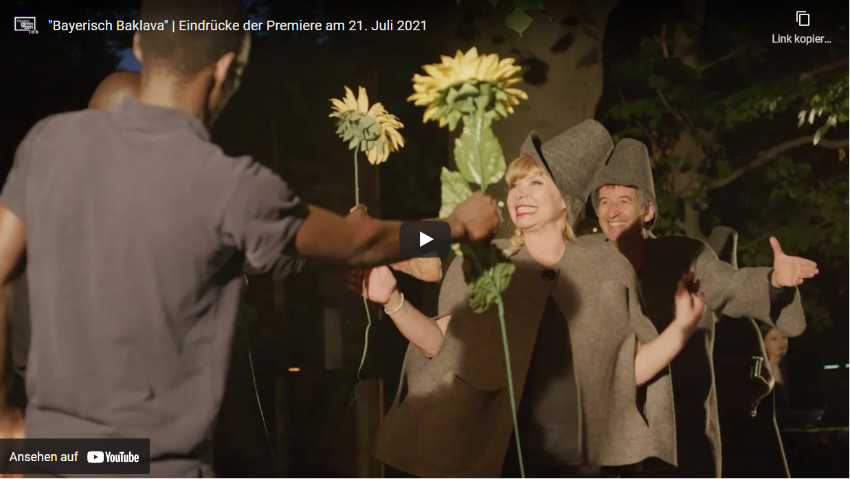 Hier sieht man einen Screenshot des YouTube-Videos über die Premiere von "Bayerisch Baklava". Darauf reicht ein junger Mann einer Dame eine Sonnenblume, die breit grinst.