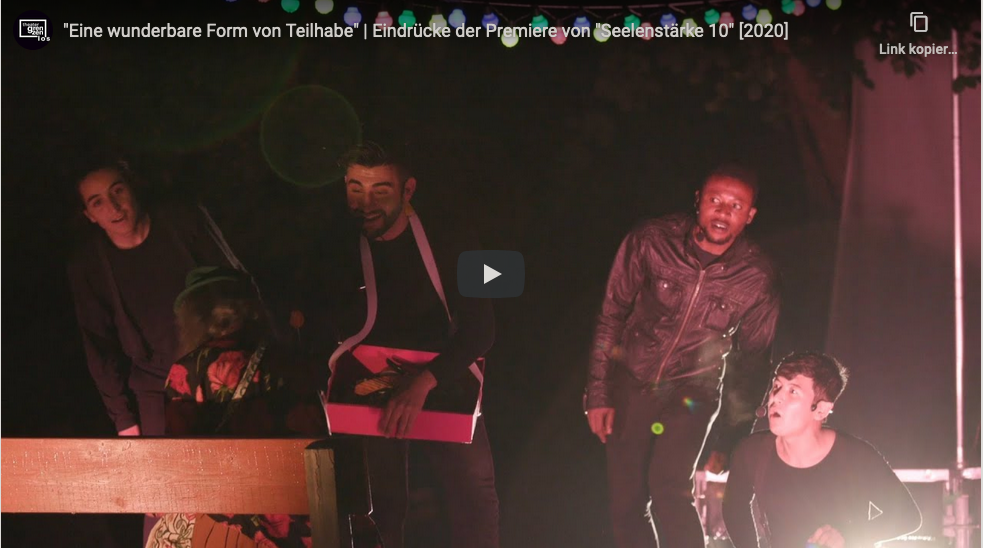 Screenshot des Teaserbildes des YouTube-Videos über die Premiere von "Seelenstärke 10", das 2020 von Theater Grenzenlos gespielt wurde. Ein junger Mann überreicht einer alten Dame eine Blume, drei andere Männer schauen gespannt zu.