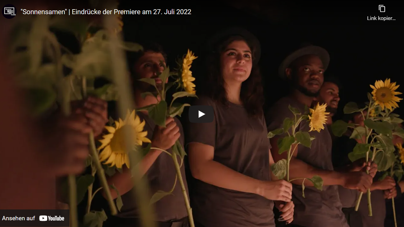 Hier sieht man einen Screenshot des YouTube-Videos über die Premiere von "Sonnensamen." Es zeigt das Ensemble mit Sonnenblumen in der Hand.