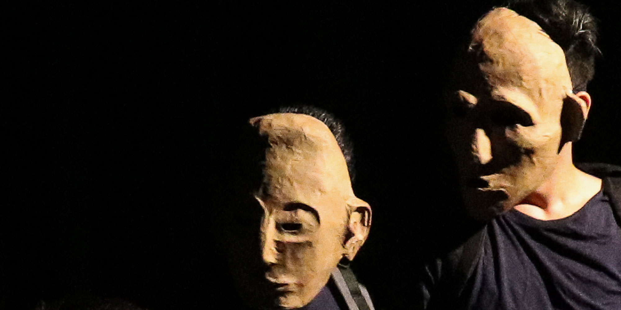 Hier sieht man zwei Schauspieler mit beigen Masken, die ihr komplettes Gesicht verdecken, die schräg nach unten blicken. Der Hintergrund ist dunkel.