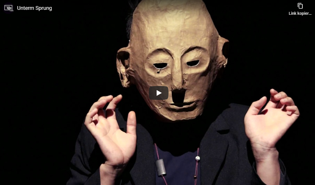 Auf dem Bild sieht man den Screenshot eines Youtube-Videos mit dem Titel "Unterm Sprung". Auf dem Foto sieht man einen Schauspieler mit einer beigen Maske frontal, der nach unten sieht und die Hände erhoben hat.