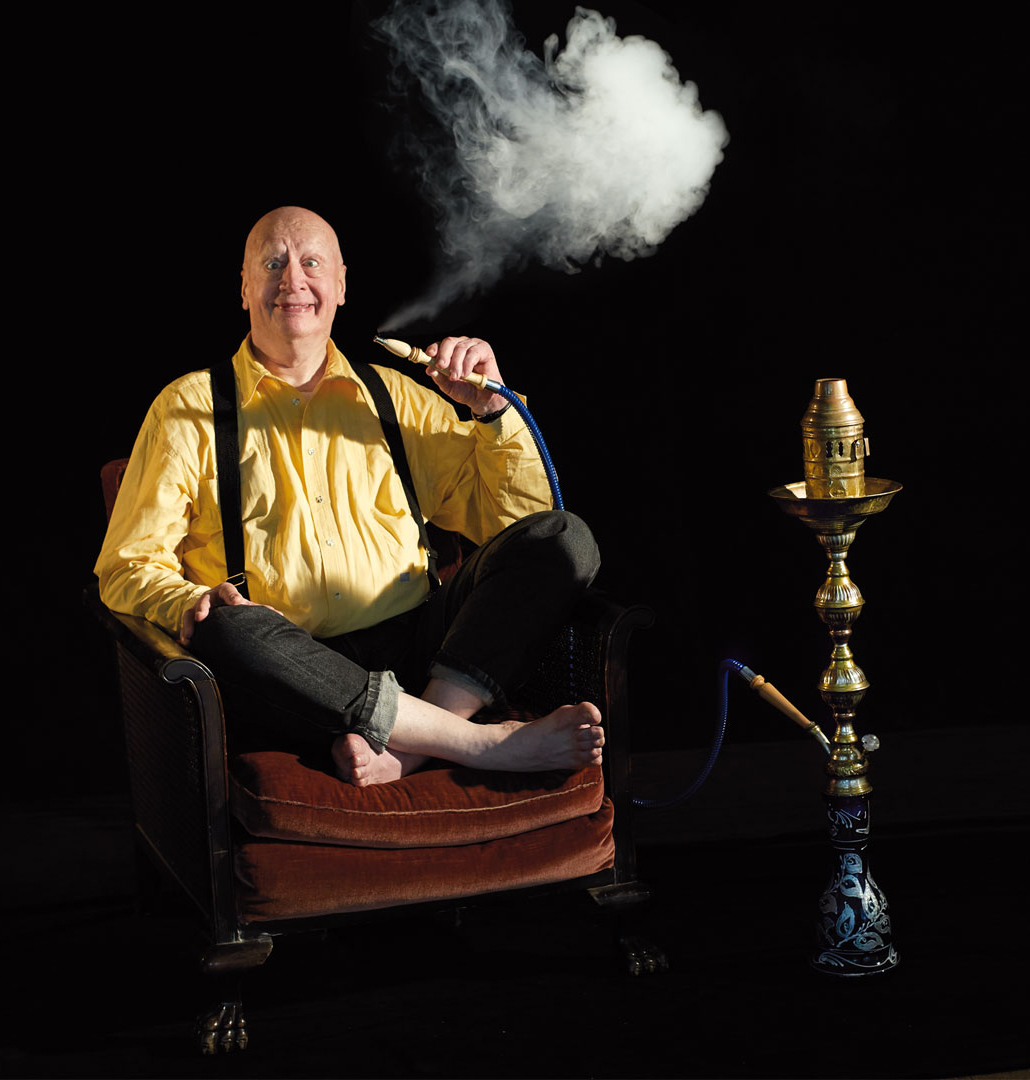 Ein älterer Mann mit Glatze sitzt im Schneidersitz auf einem Sessel und raucht Wasserpfeife. Der Hintergrund ist schwarz. Der Mann trägt ein gelbes Hemd und keine Schuhe.